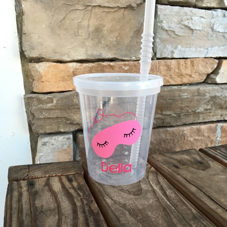 May include: A clear plastic cup with a pink sleep mask design and the name "Bella" printed on it. The cup has a straw and is sitting on a wooden surface.