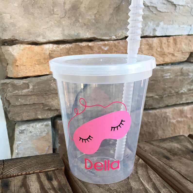 May include: A clear plastic tumbler with a pink sleep mask design and the name "Della" printed on the bottom. The tumbler has a lid and a clear plastic straw.
