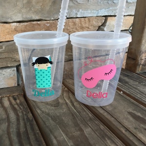 May include: Two clear plastic cups with lids and straws. The cups have personalized designs with the name "Della" and images of a girl in a sleeping bag and a pink sleep mask.