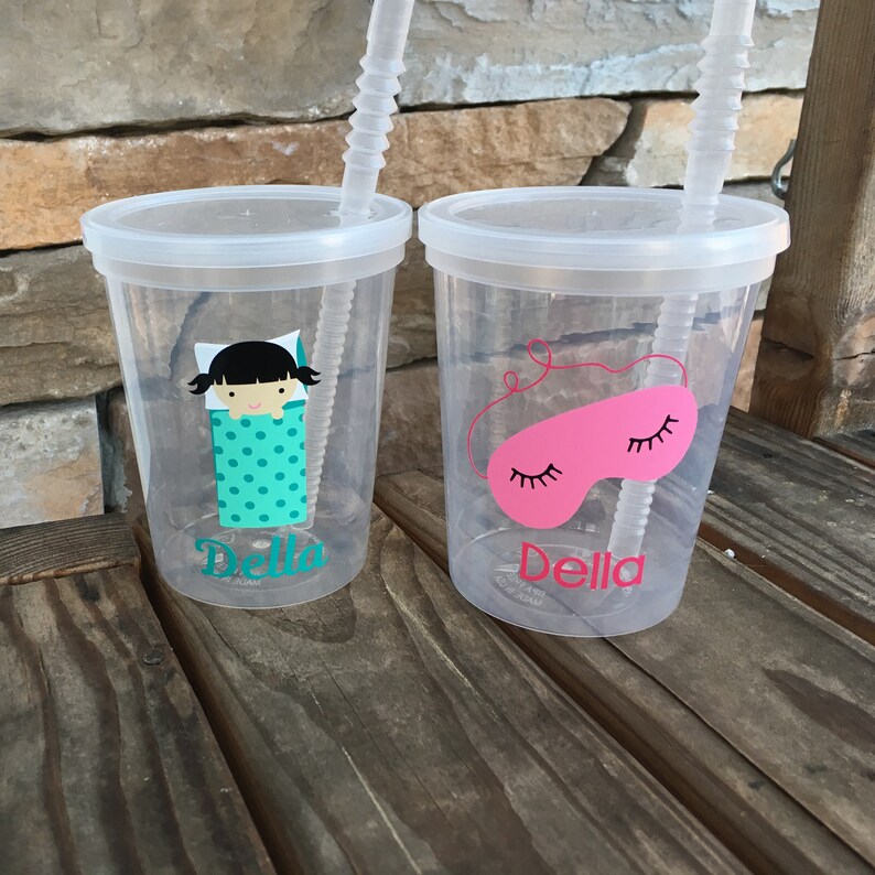 May include: Two clear plastic cups with lids and straws. The cups have personalized designs with the name "Della" and images of a girl in a sleeping bag and a pink sleep mask.