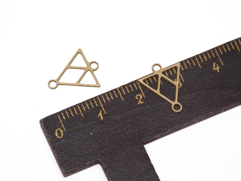 May include: Two gold-toned metal triangle charms with openwork designs and jump rings. The charms are about 1 inch in size and are shown next to a ruler.