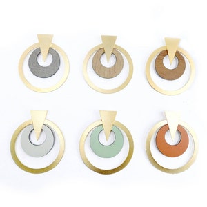 Op de afbeelding: Zes paar geometrische oorbellen. Elke oorbel heeft een goudkleurige ring met een kleinere binnenste cirkel in verschillende kleuren en texturen, waaronder grijs, houtnerf en oranje. Bovenop elke oorbel zit een gouden driehoek.
