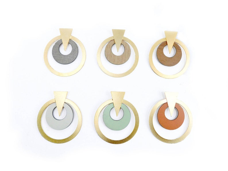 Op de afbeelding: Zes paar geometrische oorbellen. Elke oorbel heeft een goudkleurige ring met een kleinere binnenste cirkel in verschillende kleuren en texturen, waaronder grijs, houtnerf en oranje. Bovenop elke oorbel zit een gouden driehoek.