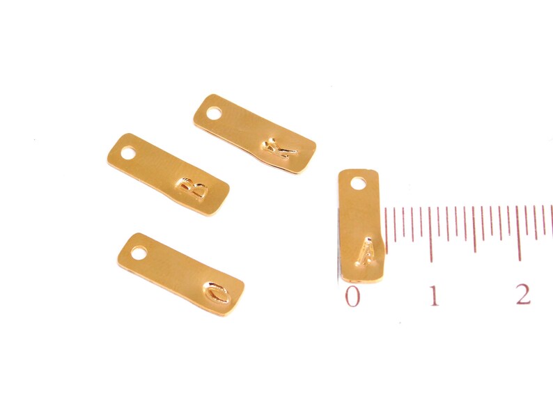 Op de afbeelding: Vier gouden metalen labels met de letters A, B en O erop gestempeld. De labels zijn ongeveer 2,5 cm lang en hebben een gat aan de bovenkant om aan een ketting of armband te bevestigen.