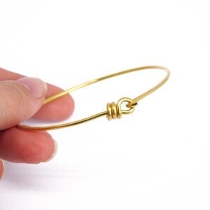 May include: A delicate gold-colored bangle bracelet. The bracelet is a simple, thin wire design with a looped clasp. The clasp is made of wrapped wire. The bracelet is held by a hand against a white background.