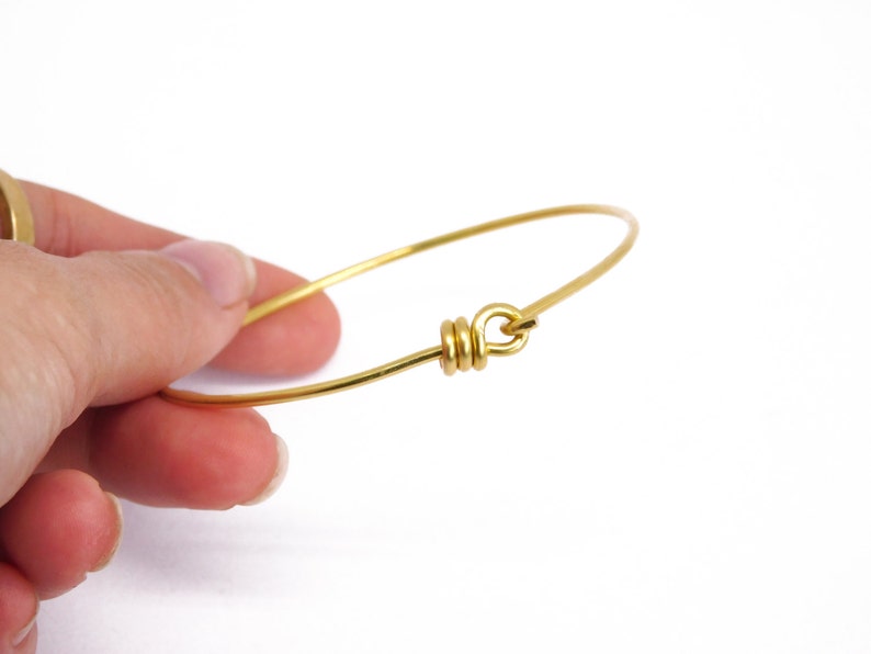 May include: A delicate gold-colored bangle bracelet. The bracelet is a simple, thin wire design with a looped clasp. The clasp is made of wrapped wire. The bracelet is held by a hand against a white background.