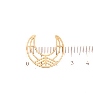 May include: A gold-colored metal crescent moon charm with a geometric design. The charm has a cutout pattern and a small hole at the top for stringing. A ruler is placed next to the charm for measurement.