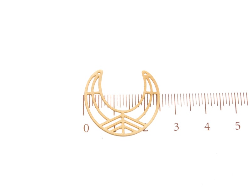 May include: A gold-colored metal crescent moon charm with a geometric design. The charm has a cutout pattern and a small hole at the top for stringing. A ruler is placed next to the charm for measurement.
