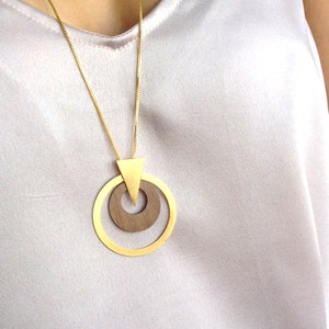Op de afbeelding: Een goudkleurige ketting met een geometrische hanger. De hanger heeft een driehoek en twee concentrische cirkels, &eacute;&eacute;n van hout en &eacute;&eacute;n van goud. De ketting wordt gepresenteerd tegen een lichtgekleurde achtergrond.