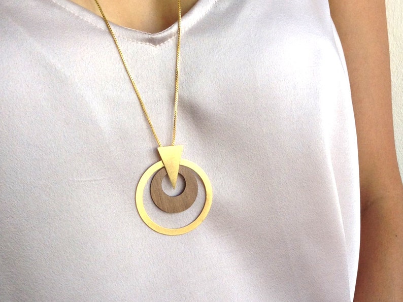 Op de afbeelding: Een goudkleurige ketting met een geometrische hanger. De hanger heeft een driehoek en twee concentrische cirkels, &eacute;&eacute;n van hout en &eacute;&eacute;n van goud. De ketting wordt gepresenteerd tegen een lichtgekleurde achtergrond.