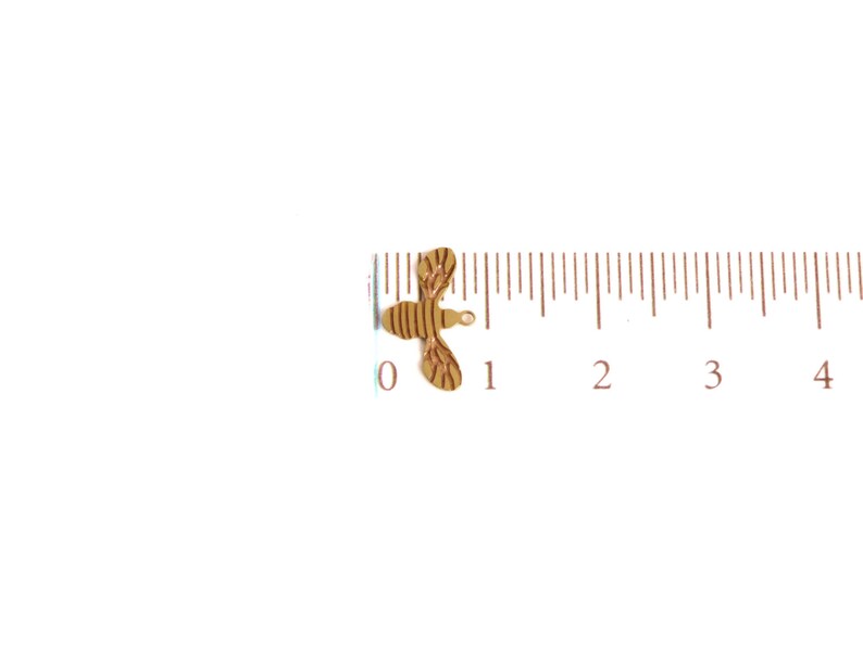 May include: A small, gold-colored bee charm with a striped pattern. The charm is about 1 inch long and is shown next to a ruler.