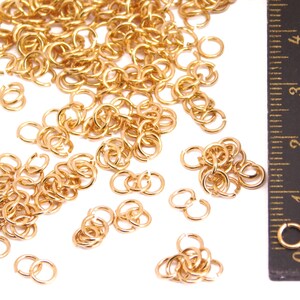 Puede incluir: Un montón grande de anillos de salto dorados, esparcidos sobre una superficie blanca. Una regla es visible en el fondo, con marcas de 0 a 4 pulgadas.