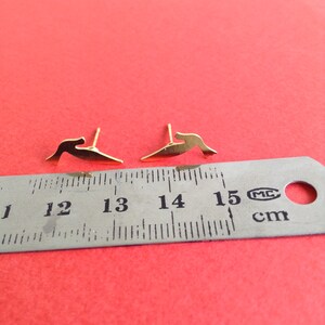 May include: A pair of gold-colored earrings shaped like a bird in flight. The earrings are measured against a ruler, showing they are approximately 1.3 cm long.