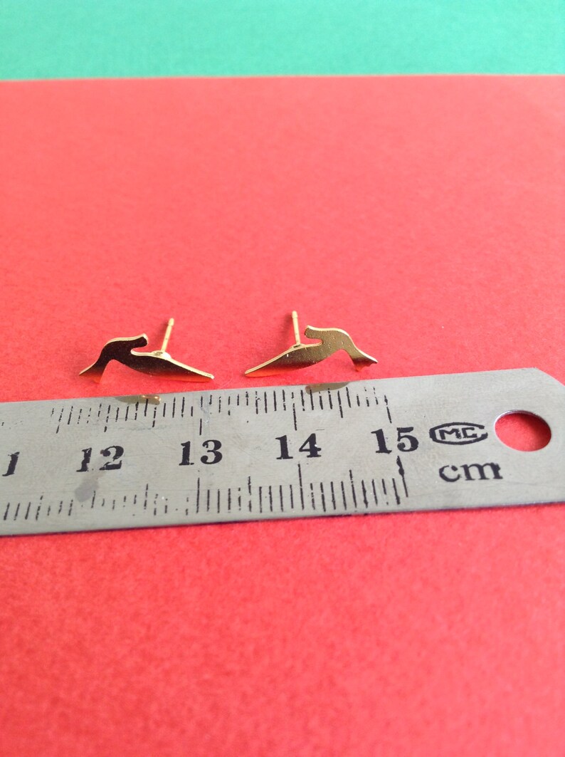 May include: A pair of gold-colored earrings shaped like a bird in flight. The earrings are measured against a ruler, showing they are approximately 1.3 cm long.