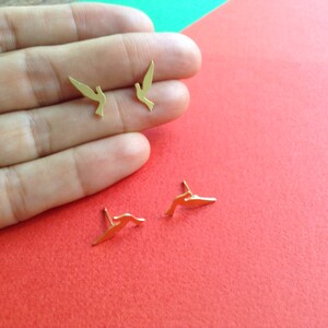 May include: A pair of gold-colored bird-shaped earrings. The earrings are small and delicate, and they are shown on a hand against a red background.