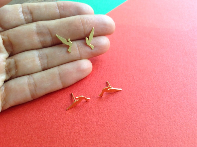 May include: A pair of gold-colored bird-shaped earrings. The earrings are small and delicate, and they are shown on a hand against a red background.