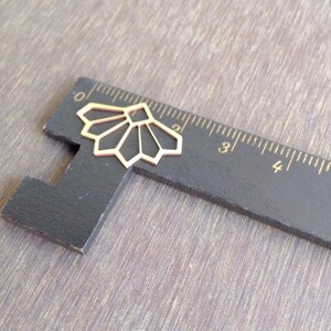May include: A gold-colored metal geometric design, resembling a fan, is placed on a black ruler with white markings. The ruler is marked with numbers from 0 to 5.