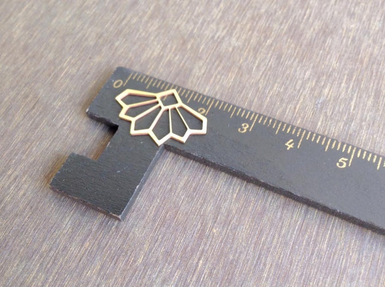 May include: A gold-colored metal geometric design, resembling a fan, is placed on a black ruler with white markings. The ruler is marked with numbers from 0 to 5.