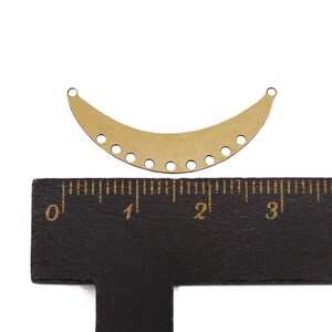 May include: A gold metal crescent-shaped pendant with a row of holes along the bottom edge. The pendant is about 2 inches wide and is sitting on a ruler.