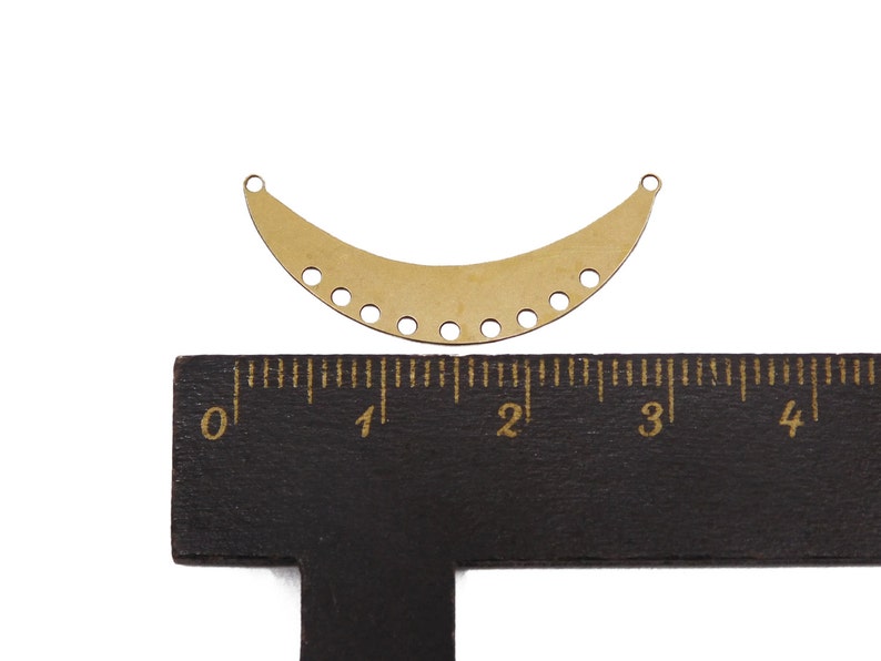 May include: A gold metal crescent-shaped pendant with a row of holes along the bottom edge. The pendant is about 2 inches wide and is sitting on a ruler.
