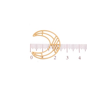 May include: A gold-colored, crescent moon-shaped charm with geometric cutouts. The charm has two small holes for attaching to jewelry. A ruler shows the charm is approximately 1 inch wide.