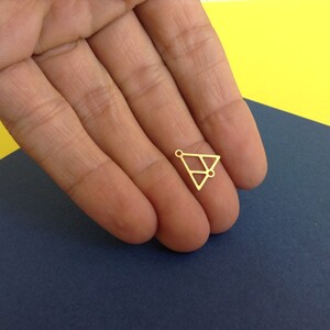 May include: Gold-toned metal triangle charm with a geometric design and three small holes. The charm is shown in a hand.