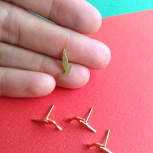 May include: Four gold-colored bird earrings. Three of the earrings are laying flat on a red surface. One earring is being held in a hand, showing the detail of the bird design.
