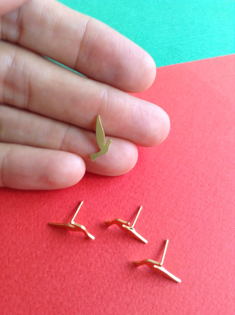 May include: Four gold-colored bird earrings. Three of the earrings are laying flat on a red surface. One earring is being held in a hand, showing the detail of the bird design.