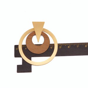 Op de afbeelding: Geometrische houten oorbellen met een goudkleurige metalen accent. De oorbellen hebben een driehoek en twee cirkels, met een donkerbruine liniaal op de achtergrond. De oorbellen zijn ongeveer 5 cm lang.