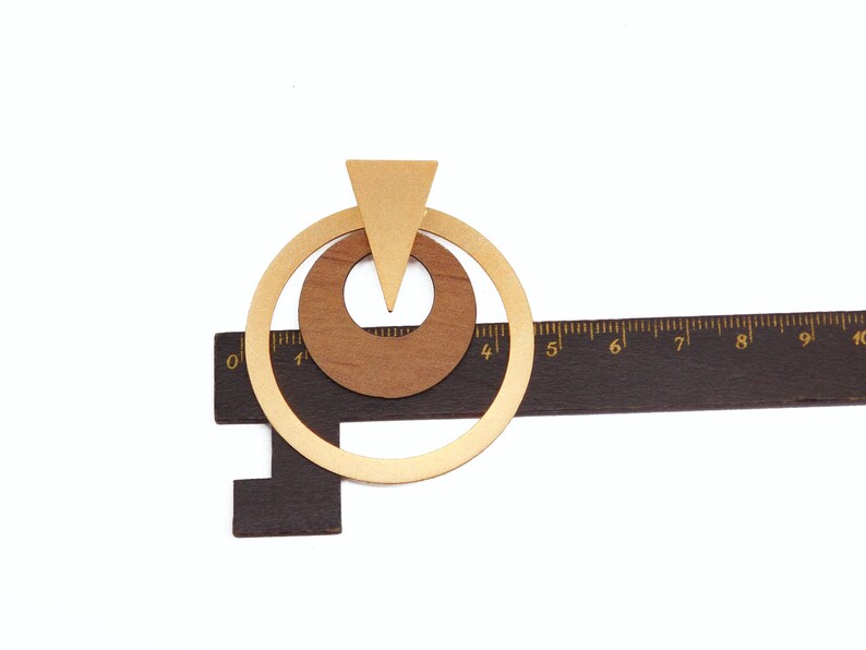 Op de afbeelding: Geometrische houten oorbellen met een goudkleurige metalen accent. De oorbellen hebben een driehoek en twee cirkels, met een donkerbruine liniaal op de achtergrond. De oorbellen zijn ongeveer 5 cm lang.