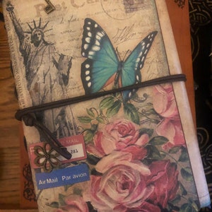 May include: A vintage-style journal with a postcard design featuring the Statue of Liberty and a large blue butterfly. The cover also displays pink roses and the words "Air Mail Par avion". A key charm and elastic band secure the journal.