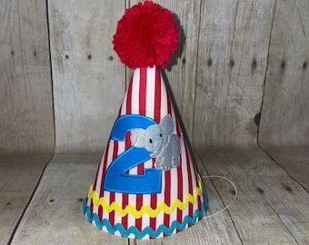 Pode incluir: Chapéu de festa listrado vermelho e branco com um número 2 azul e um elefante cinza. O chapéu tem um pompom vermelho no topo e uma borda amarela e azul.