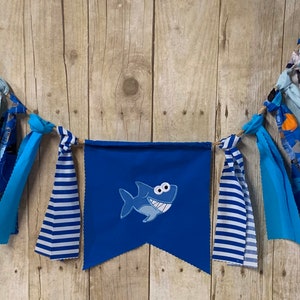 Op de afbeelding: Een blauwe en witte stoffen banner met een blauwe haai-applicatie. De banner is bespannen met blauwe, witte en grijze stoffen resten.