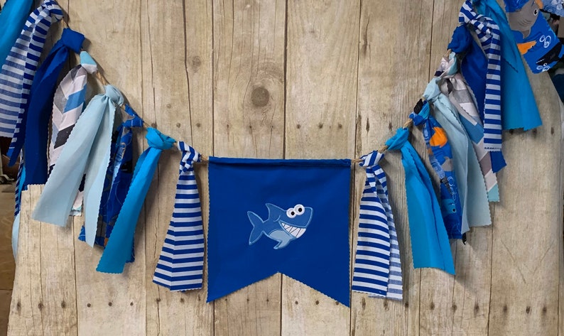 Op de afbeelding: Een blauwe en witte stoffen banner met een blauwe haai-applicatie. De banner is bespannen met blauwe, witte en grijze stoffen resten.