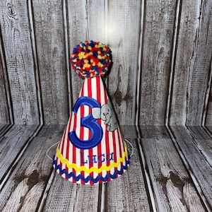 May include: Red and white striped party hat with a blue number 3, a blue elephant, and the name Luca embroidered on it. The hat has a yellow pom-pom on top.
