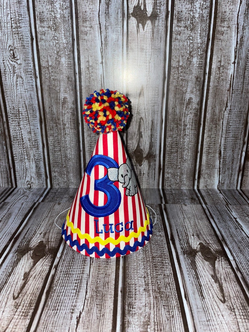 May include: Red and white striped party hat with a blue number 3, a blue elephant, and the name Luca embroidered on it. The hat has a yellow pom-pom on top.