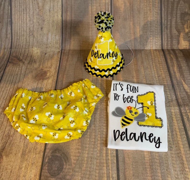 May include: A white onesie with the text "It's fun to bee" and a bee with a flower on its head. The onesie also has a large number one with bees on it. The onesie is personalized with the name "Delaney". A yellow and white diaper cover with bees on it. A yellow and black party hat with a black pom-pom on top. The hat is personalized with the name "Delaney".