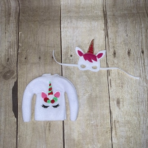 May include: White felt elf sweater with a unicorn design featuring a red, green, and white horn, pink ears, and black eyelashes. A white felt unicorn mask with a pink horn and ears is also included.