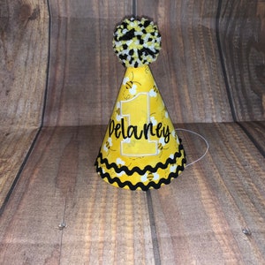 May include: A yellow and black bee-themed party hat with a white pom-pom on top. The hat has a white number "1" and the name "Delaney" embroidered on it.