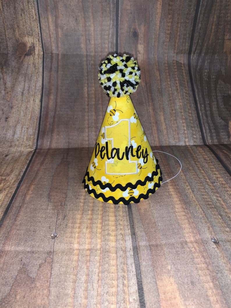 May include: A yellow and black bee-themed party hat with a white pom-pom on top. The hat has a white number "1" and the name "Delaney" embroidered on it.