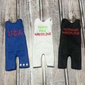 May include: Three wrestling singlets in blue, white, and black. The blue singlet has "USA" embroidered on the front and three white stars on the bottom. The white singlet has "NORTH POLE WRESTLING" embroidered on the front. The black singlet has "MUSTANGS WRESTLING" embroidered on the front.