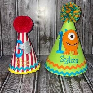 May include: Two colourful birthday party hats. One hat is red and white striped with a grey elephant and the name "Nico". The other hat is green with a one and an orange monster with the name "Sylas".