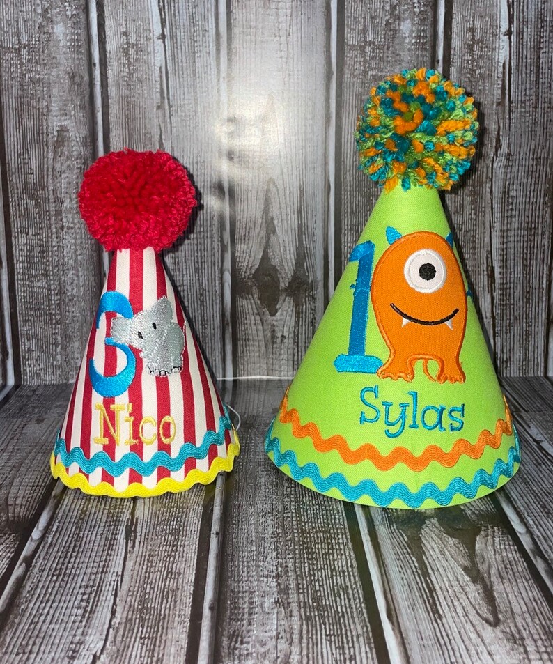 May include: Two colourful birthday party hats. One hat is red and white striped with a grey elephant and the name "Nico". The other hat is green with a one and an orange monster with the name "Sylas".