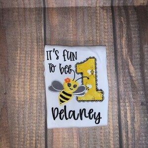 May include: White onesie with the text "It's fun to bee 1" and a bee with a yellow body, black stripes, and gray wings. The onesie also has a yellow number 1 with a lace border and the name "Delaney" in black.