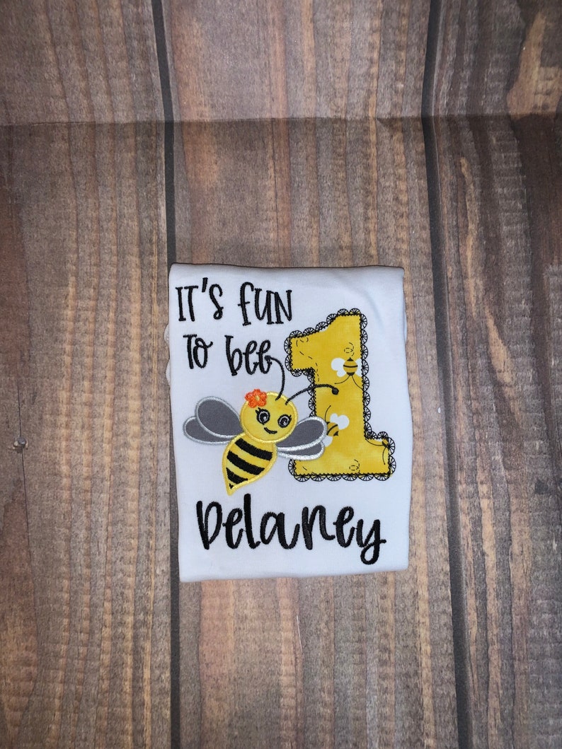 May include: White onesie with the text "It's fun to bee 1" and a bee with a yellow body, black stripes, and gray wings. The onesie also has a yellow number 1 with a lace border and the name "Delaney" in black.