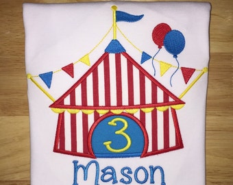 Pode incluir: Uma camiseta branca com um design de tenda de circo bordada. A tenda é listrada de vermelho e branco com uma entrada azul e amarela. O número 3 é bordado em amarelo em um círculo azul acima do texto "Mason".
