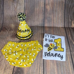 May include: A yellow and black bee-themed baby outfit with a white onesie, yellow and black diaper cover, and a yellow and black bee party hat. The onesie has the text "It's fun to bee 1 Delaney" on it.
