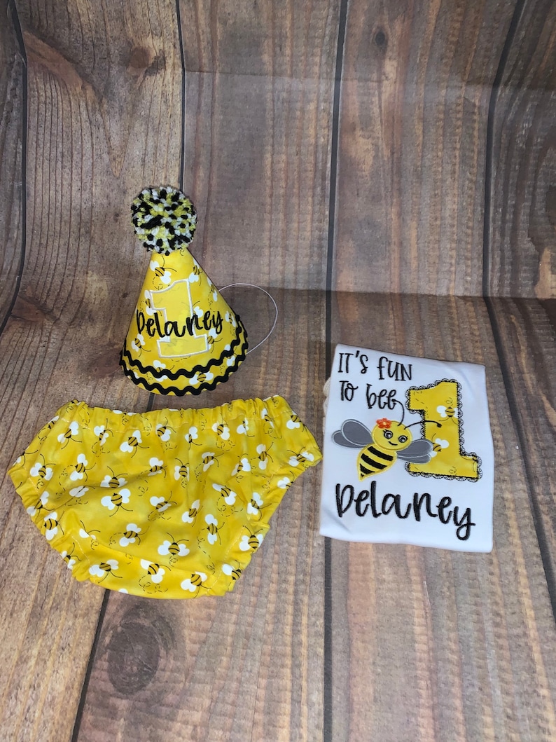 May include: A yellow and black bee-themed baby outfit with a white onesie, yellow and black diaper cover, and a yellow and black bee party hat. The onesie has the text "It's fun to bee 1 Delaney" on it.