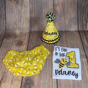 May include: A yellow and black bee-themed first birthday outfit set for a baby. The set includes a white onesie with the text "It's fun to bee" and a bee graphic, a yellow and black bee-patterned diaper cover, and a yellow party hat with the number one and the name "Delaney" on it.