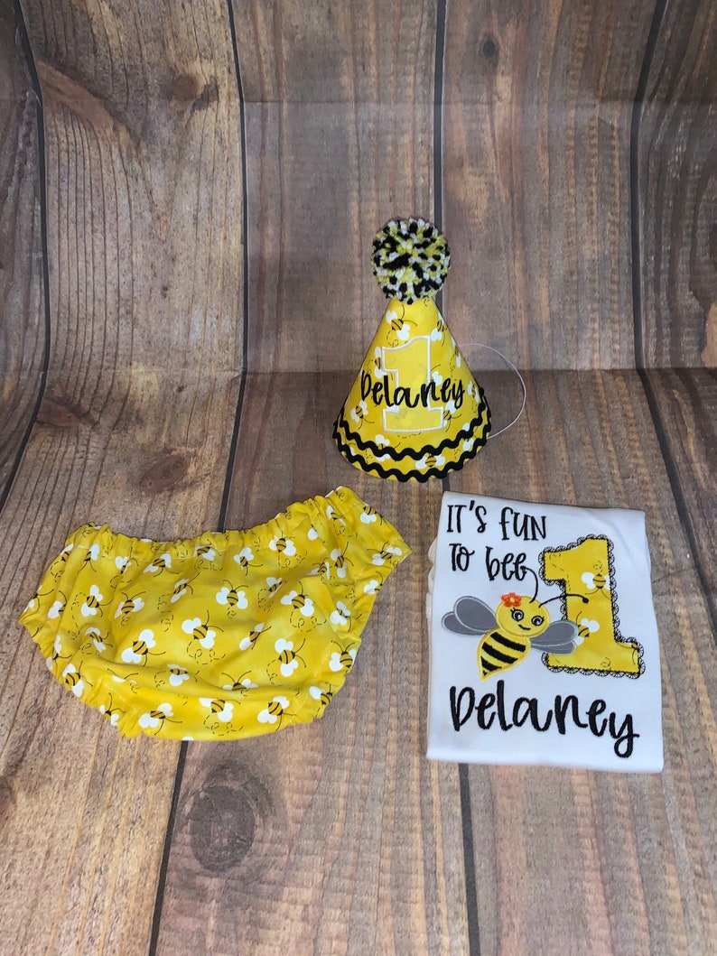 May include: A yellow and black bee-themed first birthday outfit set for a baby. The set includes a white onesie with the text "It's fun to bee" and a bee graphic, a yellow and black bee-patterned diaper cover, and a yellow party hat with the number one and the name "Delaney" on it.